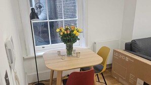 Une petite pièce moderne avec une table à manger en bois, deux chaises, un vase de fleurs, une fenêtre donnant sur un bâtiment en briques et un grand carton posé sur le sol.