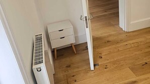 Un couloir avec un radiateur blanc, une petite table de chevet blanche et un lit dans la pièce adjacente.