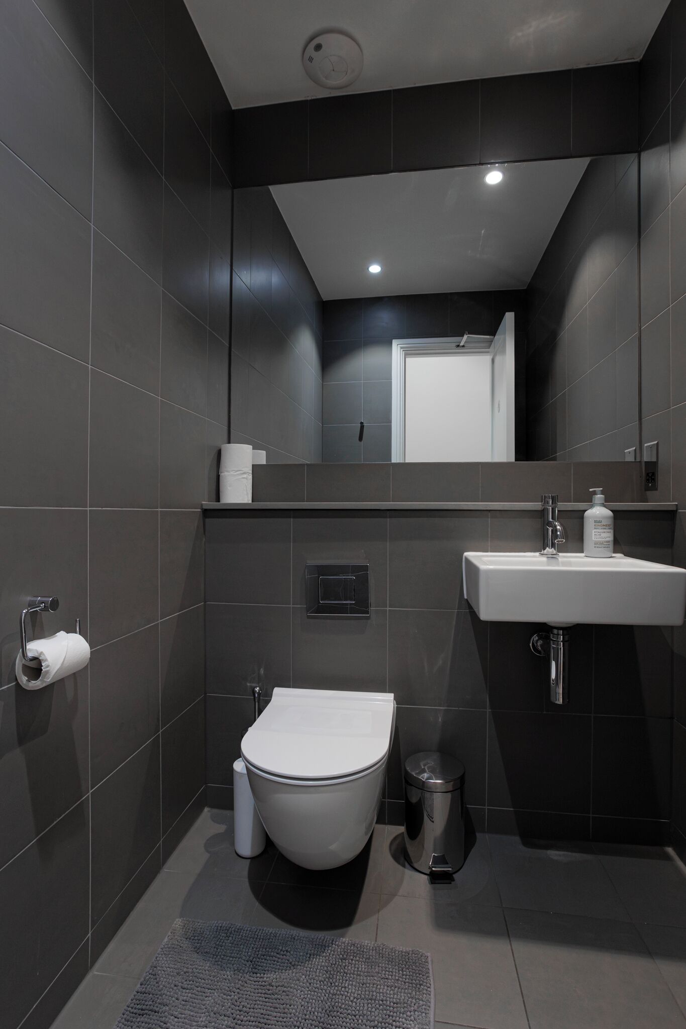 Un baño moderno con baldosas oscuras, un inodoro blanco, un lavamanos y un espejo.