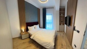 A modern hotel room with a neatly made bed, a bedside table with a lamp, a TV mounted on the wall, and a wooden wardrobe.