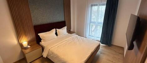 A modern hotel room with a large bed, a wooden headboard, a bedside table with a lamp, a TV, and a window with curtains.