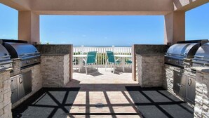An outdoor patio with two barbecue grills, a white fence, and a view of the ocean.
