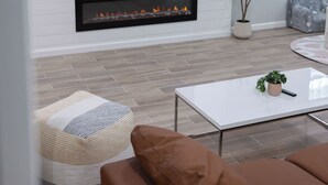 A modern living room with a flat-screen TV mounted on a white wall, a fireplace below, and a comfortable seating area with a brown sofa and a white coffee table.