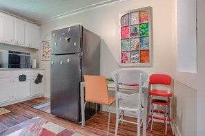 A compact kitchen with white cabinets, a stainless steel refrigerator, a microwave, and a colorful window. There are two chairs, one orange and one red, and a small table.