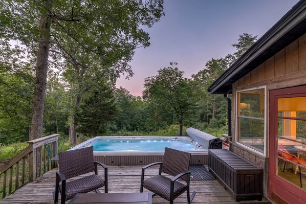 A hot tub on a wooden deck with wicker chairs, surrounded by trees.