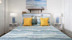 Un dormitorio con una cama, dos mesitas de noche, un estante en la pared y un cuadro de una playa.