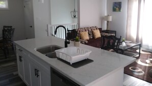 A modern kitchen with a white countertop, a sink, and a built-in oven. There is a dining area with a table and chairs, and a living room with a sofa and a lamp. - Relax and enjoy your home away from home. (Strathroy)