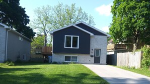 A two-story house with a dark exterior and light brick accents, a front yard with a well-maintained lawn, and a driveway leading to the front door. - Relax and enjoy your home away from home. (Strathroy)