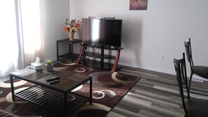 A living room with a TV on a stand, a coffee table, a chair, a small table with a vase of flowers, and a painting on the wall. - Relax and enjoy your home away from home. (Strathroy)