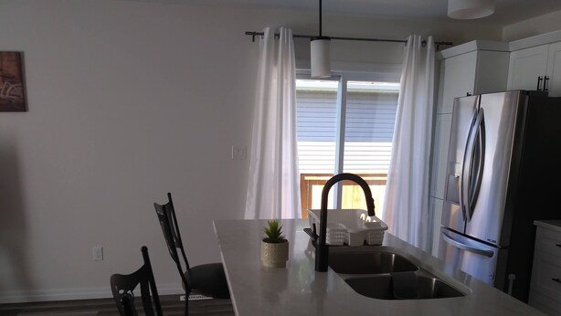A modern kitchen with a central island, stainless steel appliances, and a window with curtains. - Relax and enjoy your home away from home. (Strathroy)