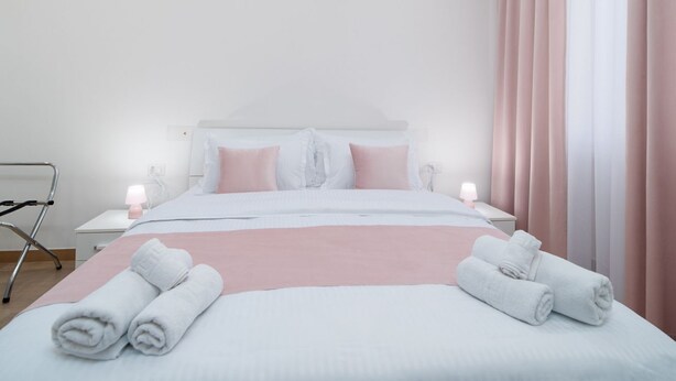 A hotel room with a bed, pillows, a bedside table with a lamp, and a window with curtains. - Lovely Rooms by Rentbeat (Civitavecchia)