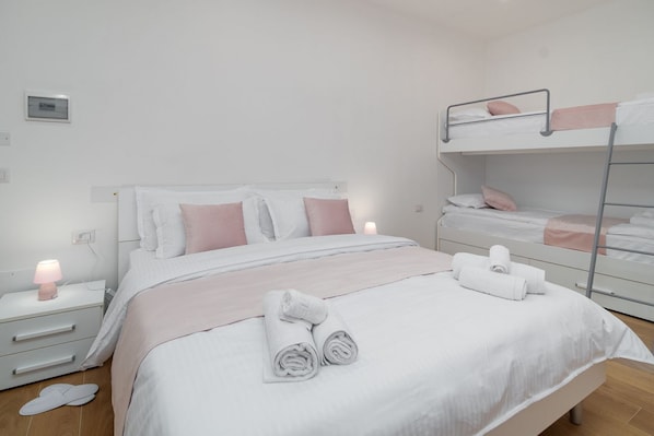 Ein ordentlich bezogenes Bett in Weiß und Rosa, ein Nachttisch mit Lampe sowie ein Etagenbett zur Rechten.