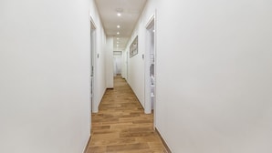 A narrow hallway with white walls and wooden flooring, leading to a door at the end.