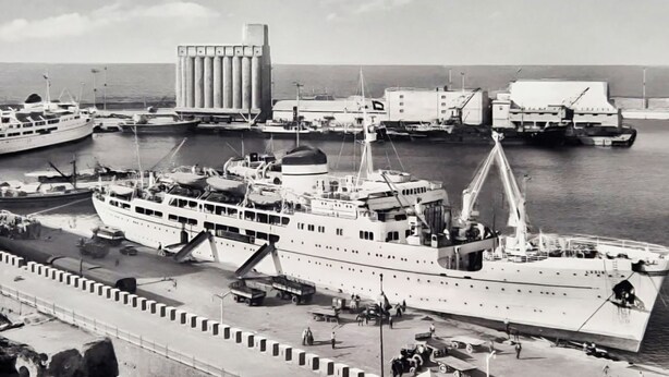 A large ship docked at a port with multiple smaller boats and a tall building in the background.