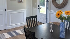 Un comedor con una mesa de madera oscura, dos sillas, un jarrón con girasoles y un espejo tejido en la pared.