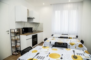 Une cuisine moderne avec des meubles blancs, un four à micro-ondes et une cuisinière. On y trouve également un coin repas avec des tables recouvertes d’une nappe aux motifs culinaires.