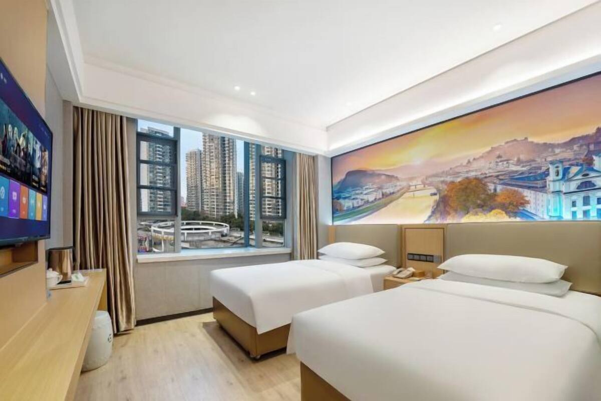 Habitación de hotel con dos camas, un ventanal grande con vista a la ciudad, un cuadro en la pared y un televisor a la izquierda.