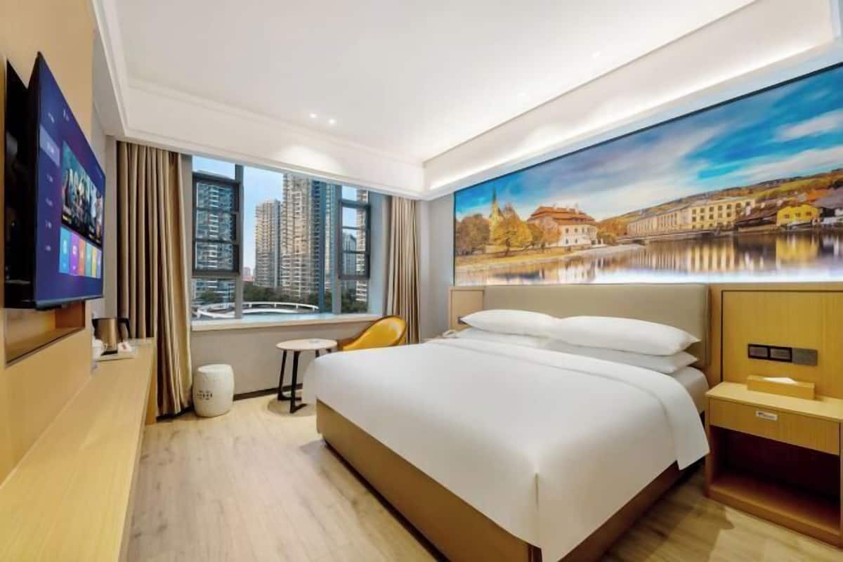 Habitación de hotel con una cama grande, televisión, una mesita, una silla y un cuadro de un paisaje urbano.