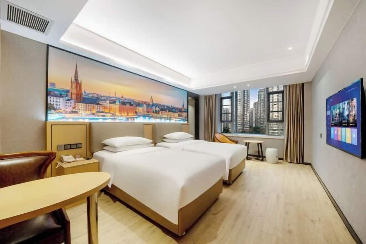 Habitación de hotel con dos camas, un escritorio, una silla, un televisor y un cuadro con una vista urbana.