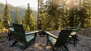 Two Adirondack chairs facing each other on a gravel surface with a forest and mountains in the background.