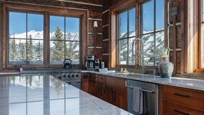 Una cocina amplia con una isla grande, armarios de madera y ventanas panorámicas con vistas a montañas nevadas.