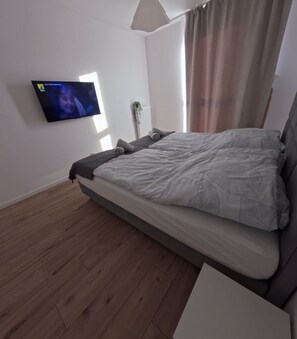 A modern bedroom with a large bed, a flat-screen TV mounted on the wall, and a window with curtains. - Stylish seaside escape in Nowe Pogórze! Relax & enjoy the view! Wifi free (Pogórze)