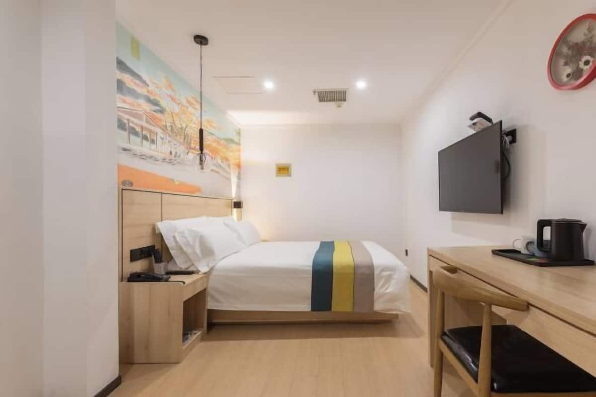 A hotel room with a bed, bedside table, desk, chair, TV, and artwork on the wall.
