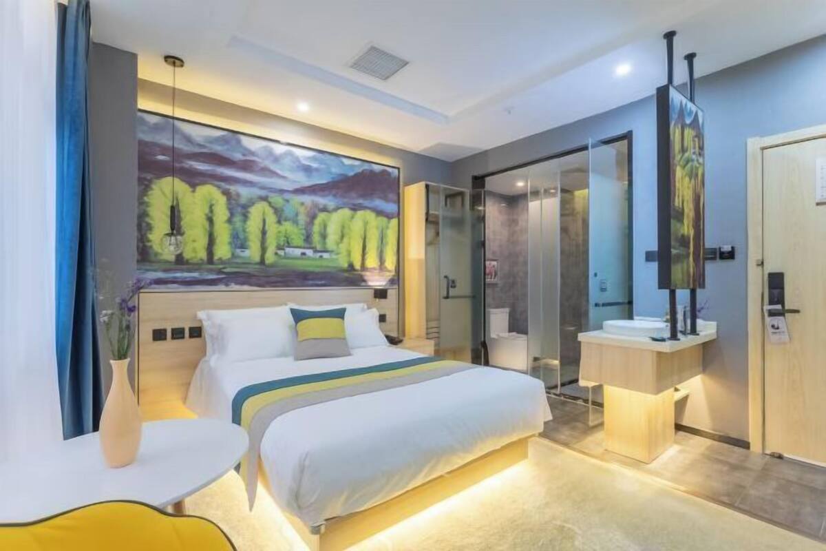 A modern hotel room with a large bed, bedside tables, a glass shower enclosure, and a large landscape painting on the wall.