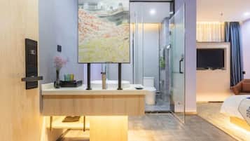 A modern hotel room with a wooden vanity, a large painting, a glass shower enclosure, and a bed with a headboard.