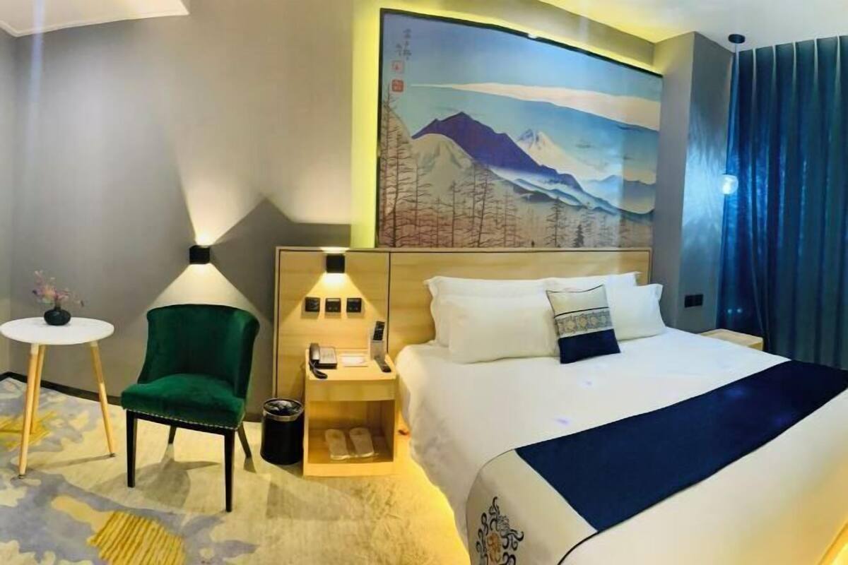 A hotel room with a bed, a green chair, a small table, a nightstand, and a painting of mountains.