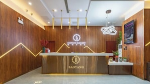 A hotel reception desk with a wooden backdrop, a logo, and a modern light fixture. - Baoyang Boutique Hotel (Shijiazhuang North Zhonghua Street Zhongchu Plaza) (Shijiazhuang)