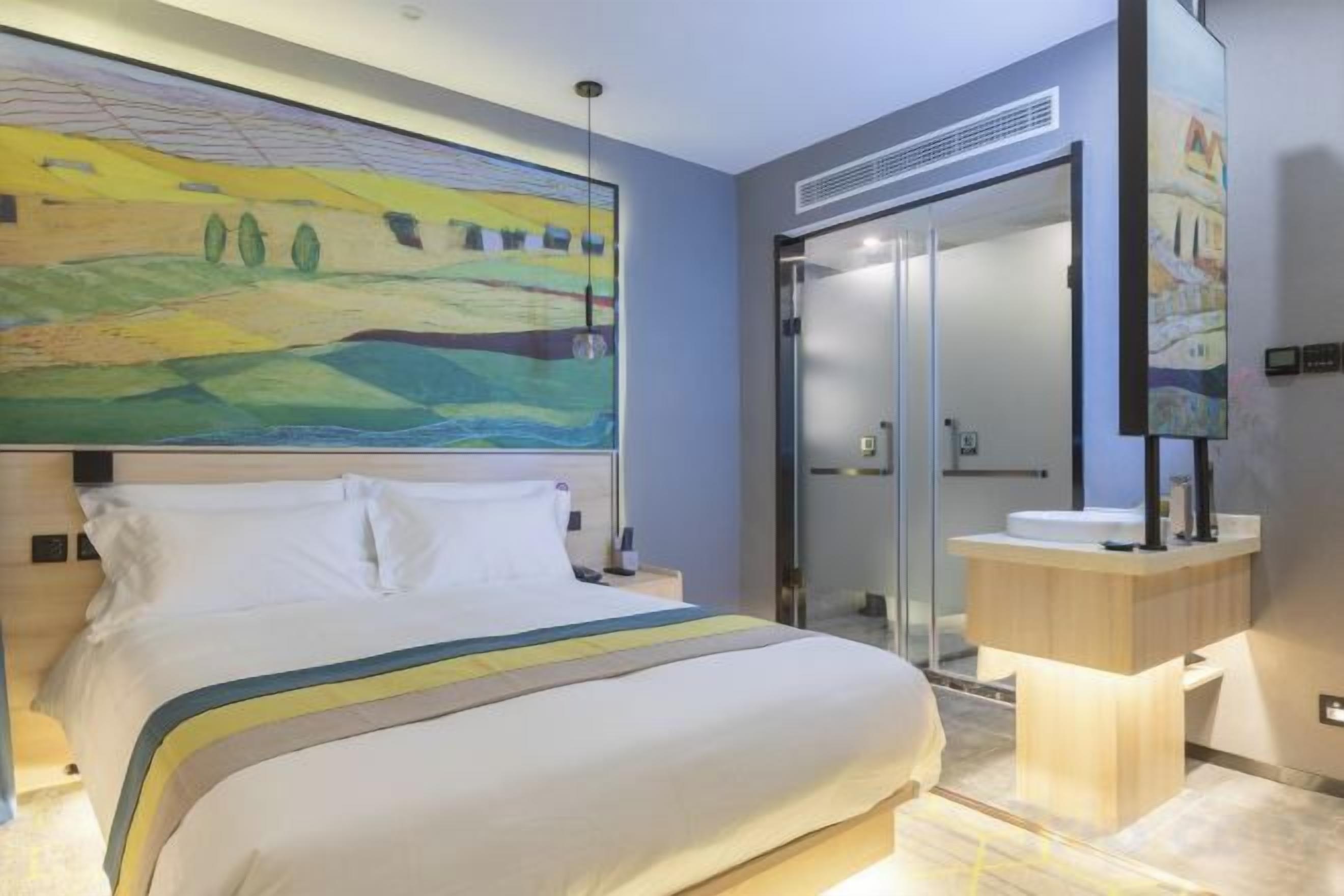Une chambre d’hôtel avec un grand lit, une table de chevet, une commode, une salle de bain avec un lavabo et un grand tableau au-dessus du lit.