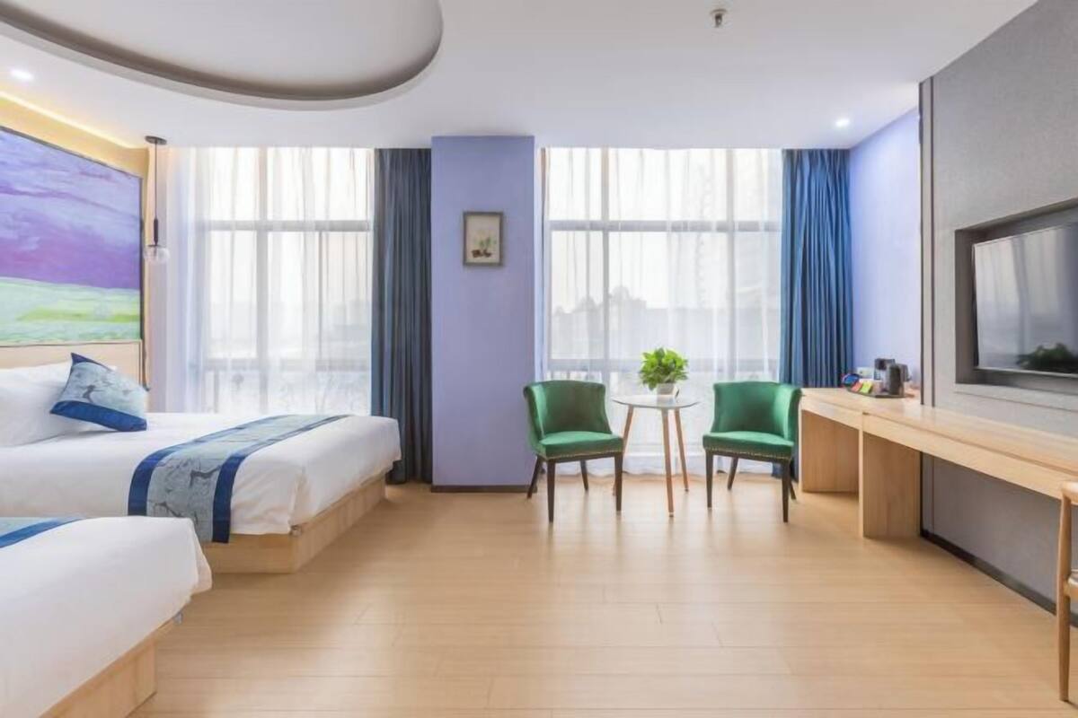A hotel room with two beds, a desk, two chairs, a TV, and large windows.