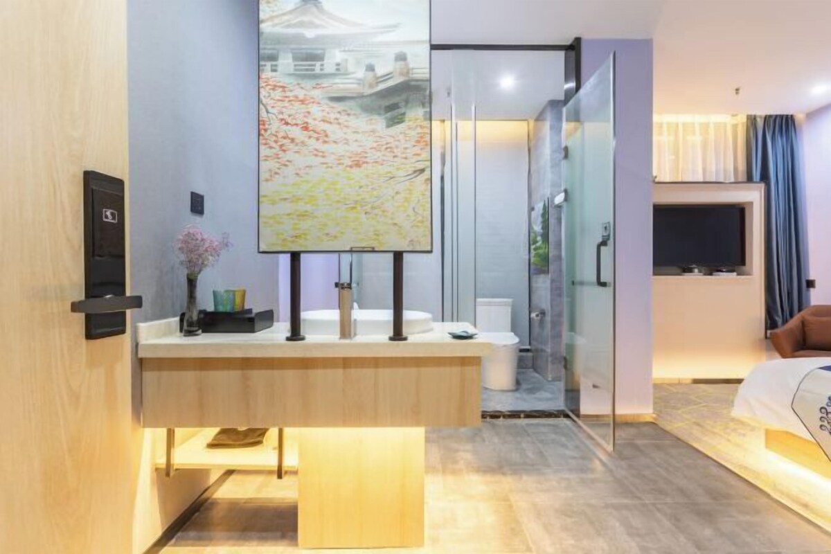 A modern hotel room with a wooden vanity, a large painting, a glass shower enclosure, and a bed with a headboard.