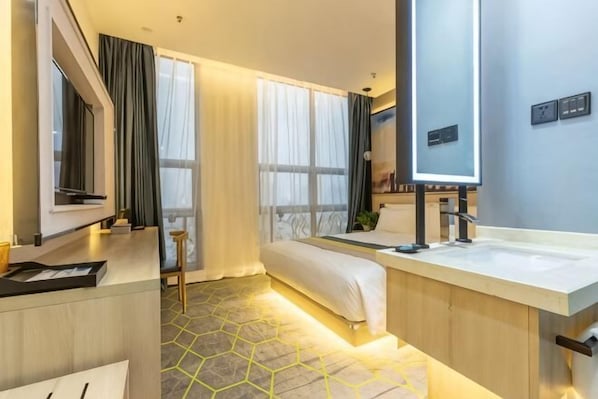 A modern hotel room with a bed, a desk, a large mirror, a sink, and a window with curtains.