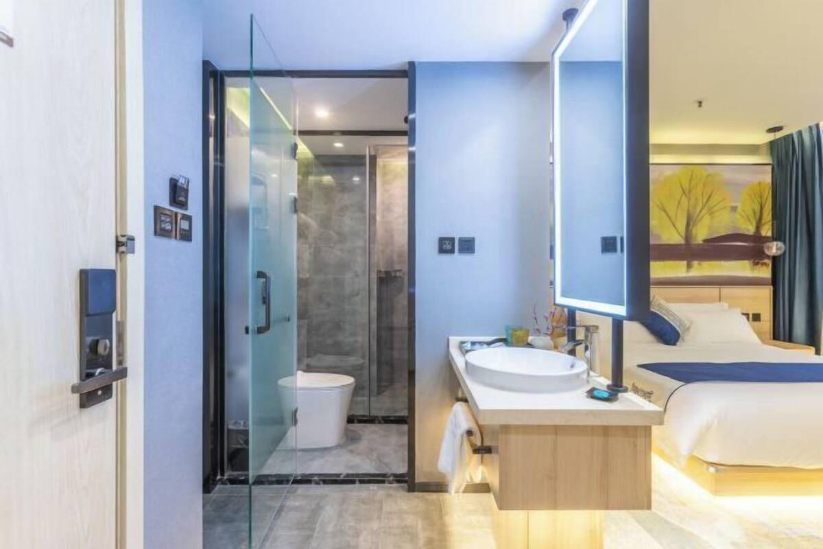 A modern hotel room with a glass-enclosed shower, a wooden vanity with a sink, and a bed with a blue headboard.