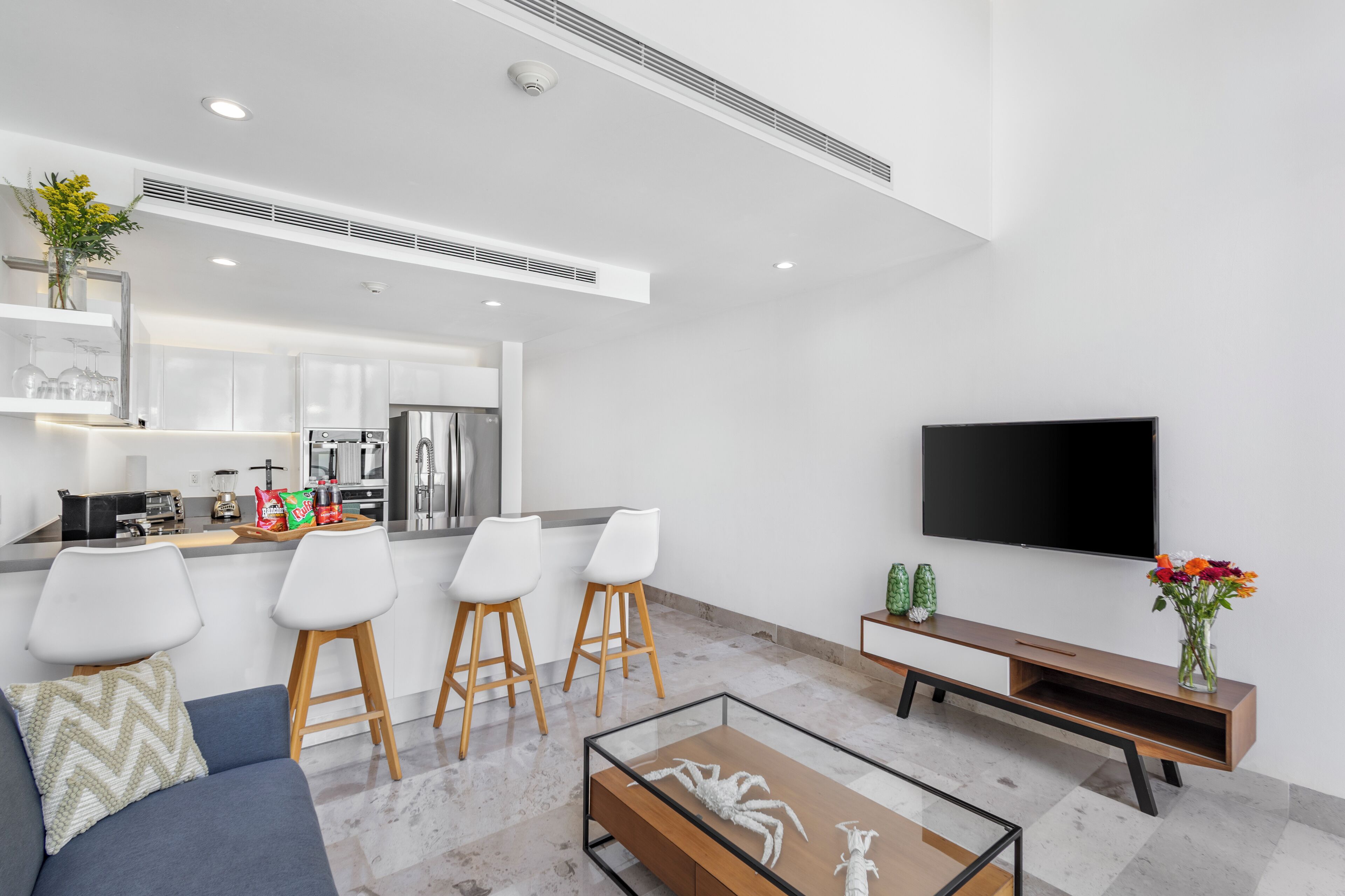 Una cocina moderna con armarios blancos, horno empotrado y refrigerador. Hay tres taburetes blancos y un banco de madera. Un televisor de pantalla plana está montado en la pared.