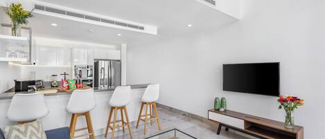 Una cocina moderna con armarios blancos, horno empotrado y refrigerador. Hay tres taburetes blancos y un banco de madera. Un televisor de pantalla plana está montado en la pared.