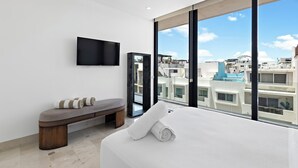 Une chambre d’hôtel moderne dotée d’un grand lit, d’une banquette, d’un téléviseur à écran plat et offrant une vue sur la ville grâce à de grandes fenêtres.