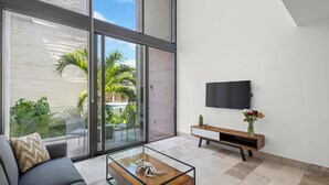 Un salon moderne avec une table basse en verre, un canapé, un téléviseur fixé au mur et une porte-fenêtre coulissante donnant sur un espace extérieur agrémenté de plantes.