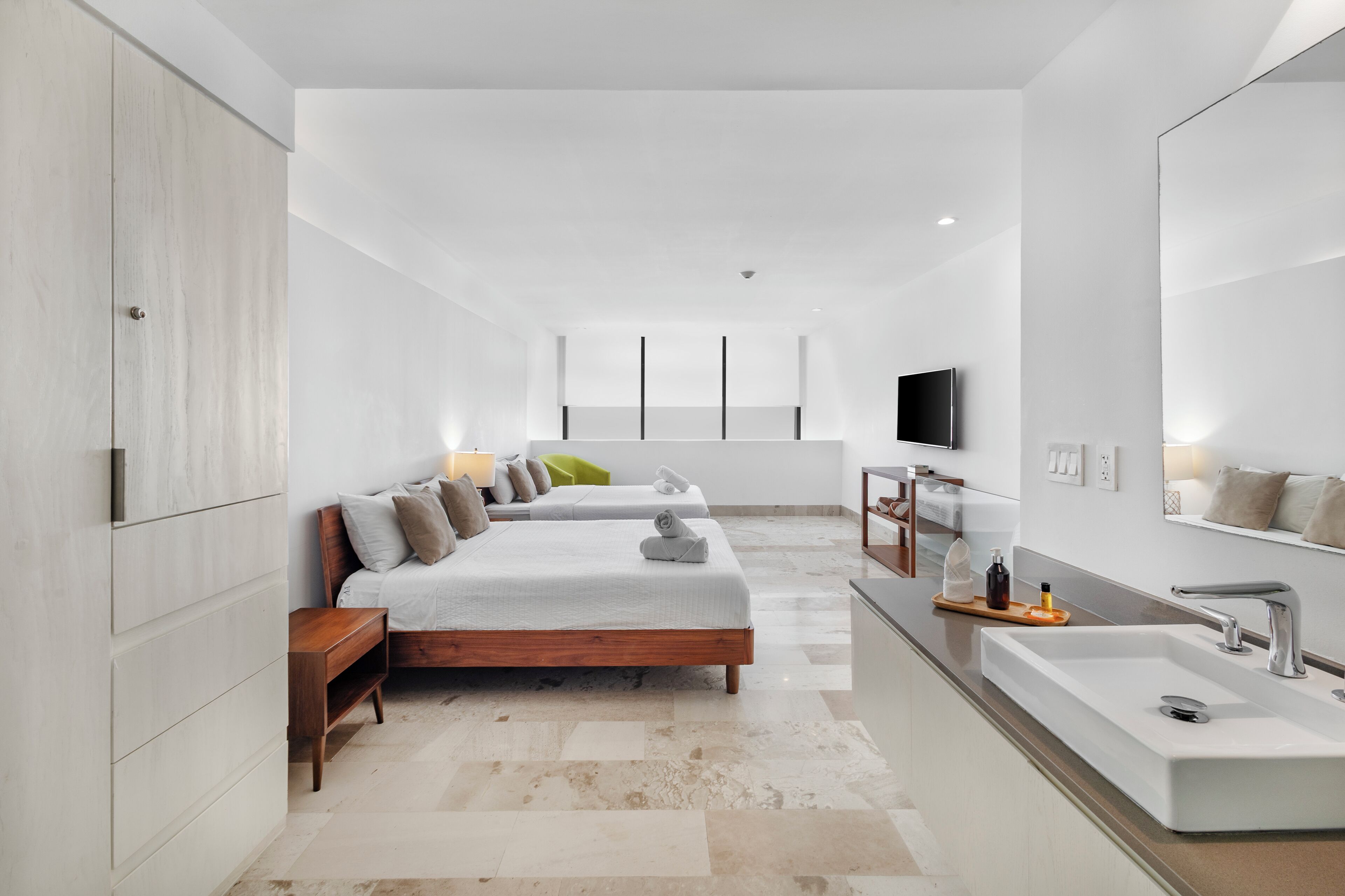 A modern hotel room with a large bed, a sitting area, a bathroom with a large mirror, and a TV mounted on the wall.