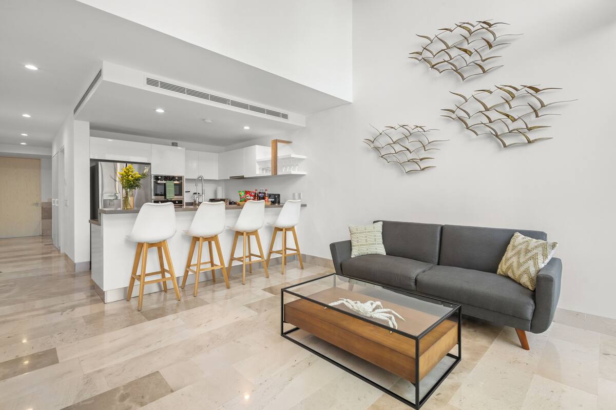 Een moderne keuken met witte kasten, een centraal eiland met barstoelen, en een grijze bank met een glazen salontafel in de woonkamer.