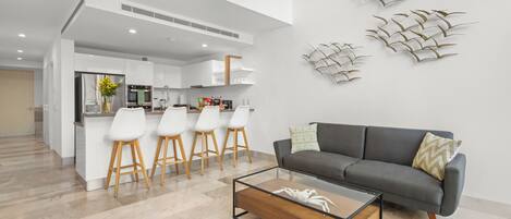 A modern kitchen with white cabinets, a central island with bar stools, and a grey sofa with a glass coffee table in the living area.