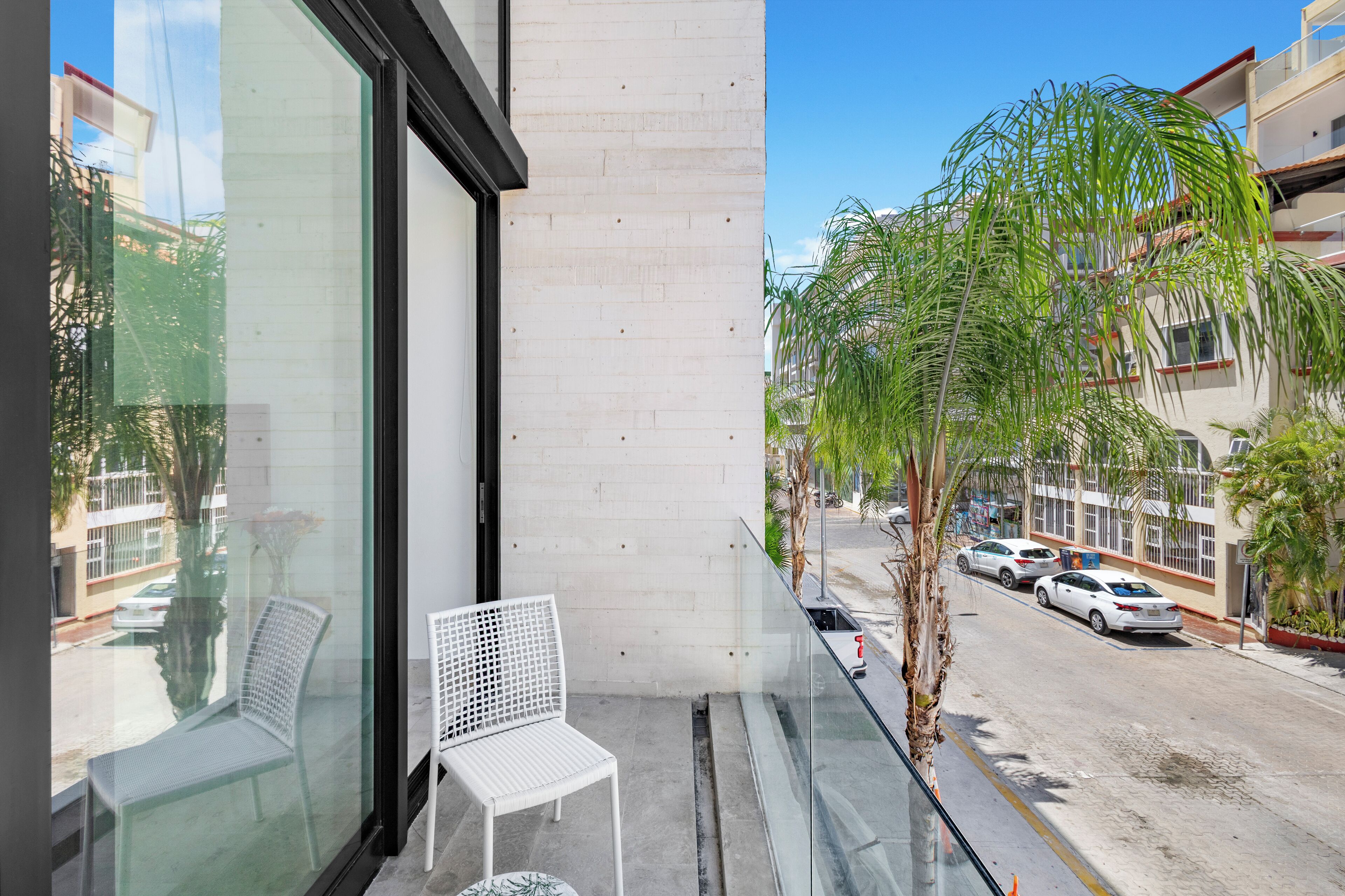 A balcony with two white chairs, a glass table, and a view of a street with parked cars and palm trees.