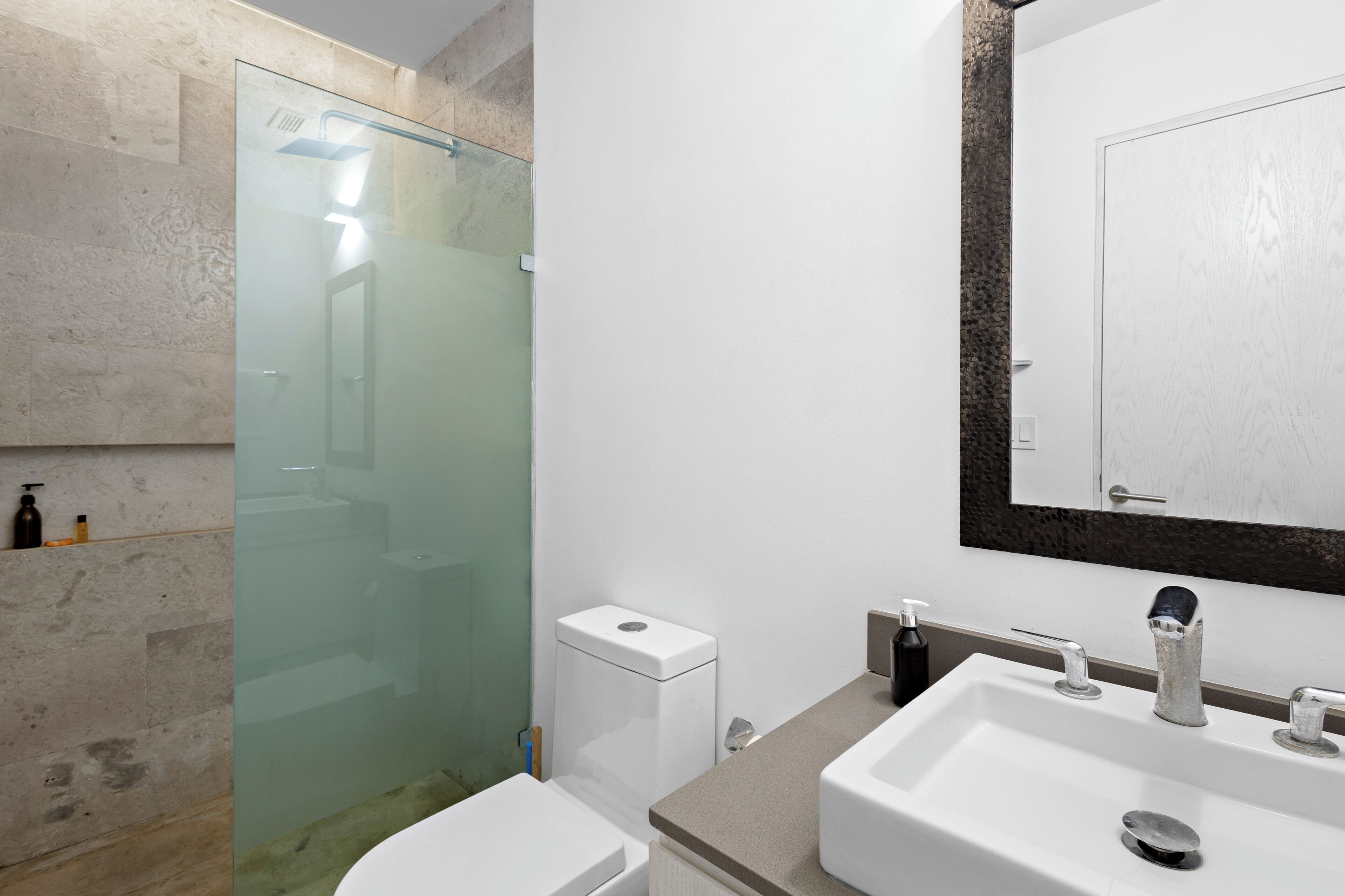 A modern bathroom with a glass shower enclosure, a white toilet, a sink with a mirror above it, and a soap dispenser on the wall.
