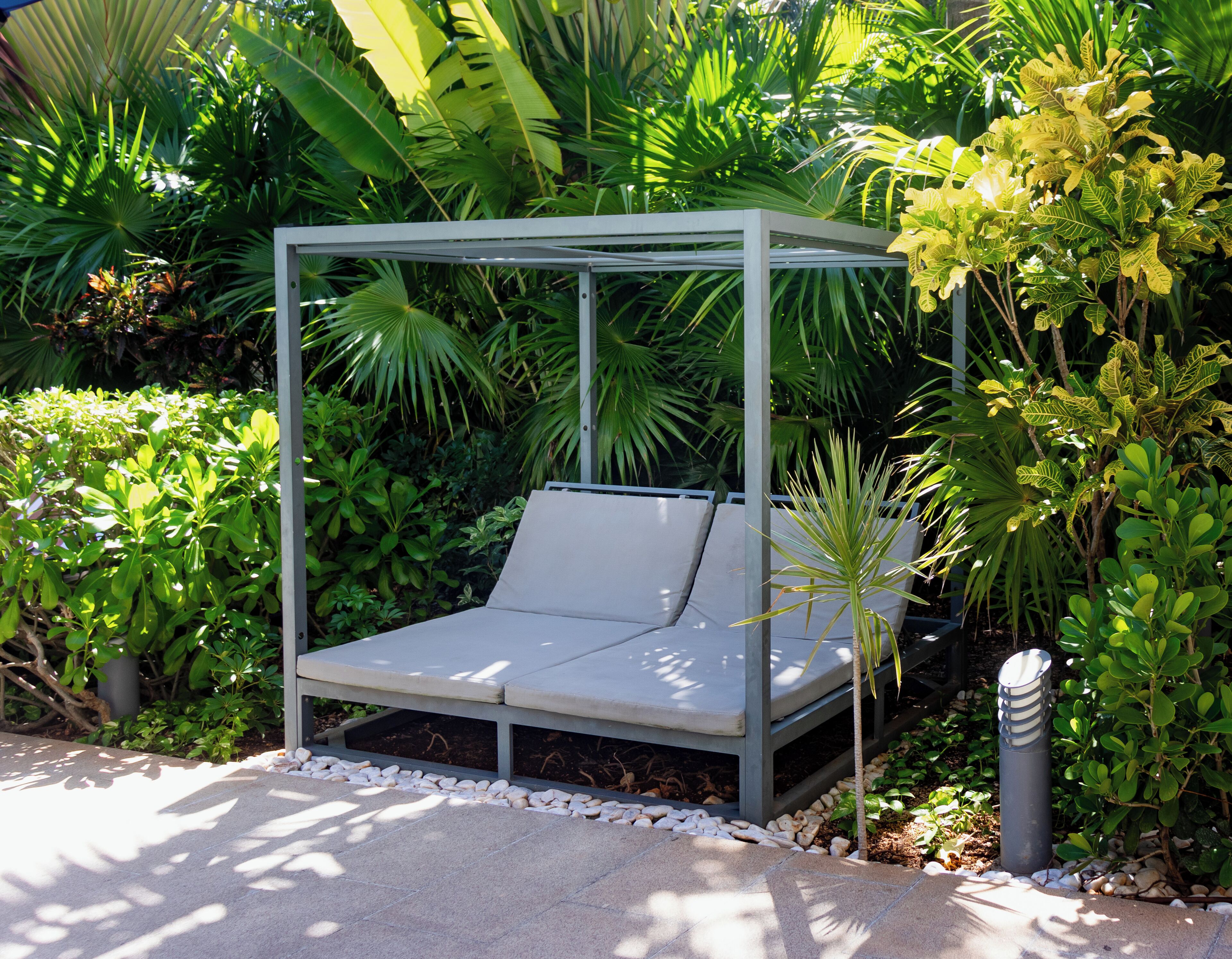 A metal gazebo with a cushioned bench, surrounded by lush greenery and plants.