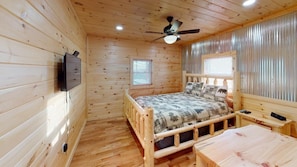 Quarto de cabana de madeira com cama, uma mesinha, um ventilador de teto e televisão fixada na parede.