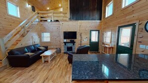 Interior de una cabaña de madera con sala de estar, una cocina con encimeras de granito, chimenea y una escalera que sube a un nivel superior.