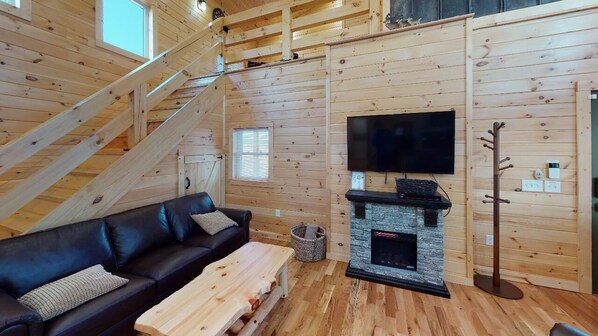A cozy cabin interior with a wooden staircase, a black leather sofa, a wooden coffee table, a stone fireplace, and a mounted television.