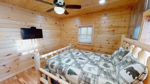 A wooden cabin room with a bed, a ceiling fan, a mounted TV, and a window.